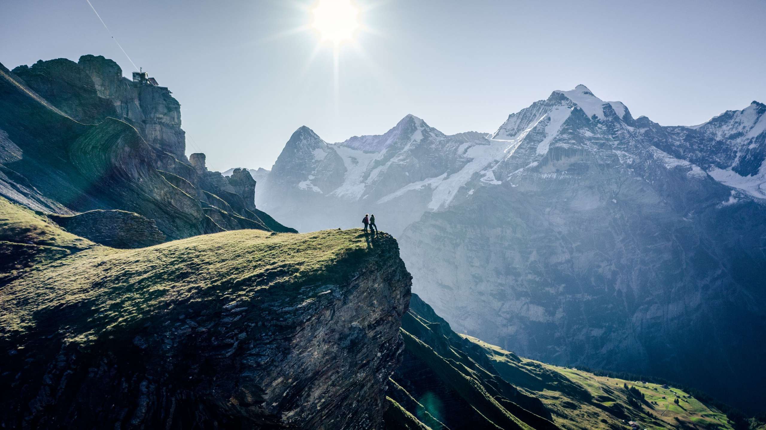 Zwei Wanderer geniessen die Aussicht vom Grauseeli auf einer Klippe. Die Station Birg und das Dreigestirn Eiger, Mönch und Jungfrau strahlen um die Wette.
